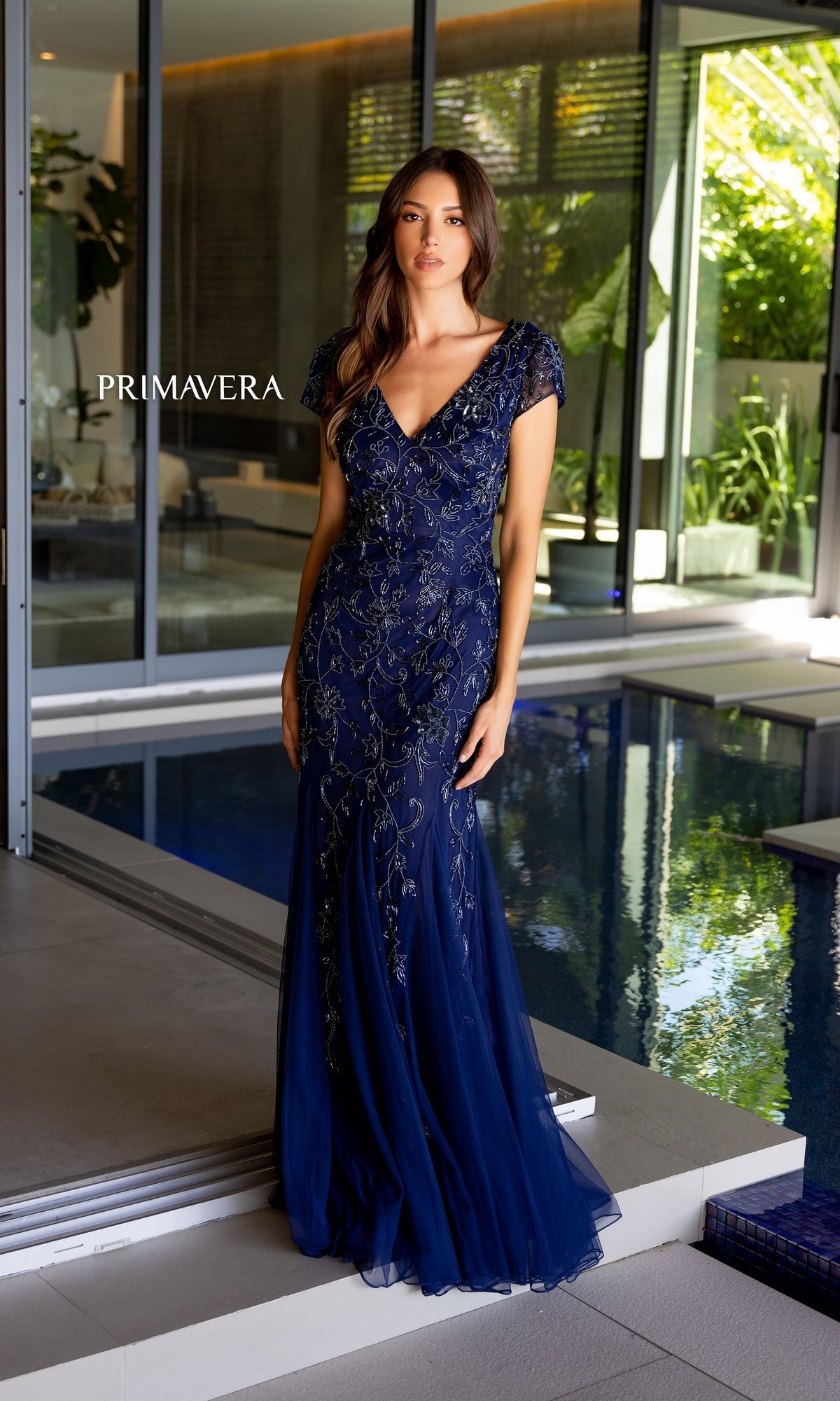 A woman stands by an indoor pool wearing the Long Prom Dress 13112 by Primavera, a navy blue floor-length mermaid gown with intricate floral beaded detailing. The word